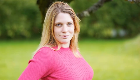 Woman in bright pink ribbed sweater poses outdoors against blurred green background.