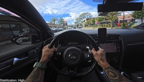 Driver's perspective from inside a car with hands on steering wheel, looking out at a sunny street with parked vehicles and brick buildings.