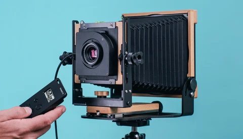 Large format camera with wooden frame and bellows, held alongside a cable release against a blue background.