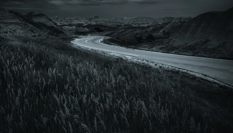 Winding road cutting through dense coniferous forest with mountains in the distance under dark, overcast sky.