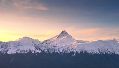 Snow-covered mountain peaks lit by golden sunrise light, with forested slopes in silhouette below.