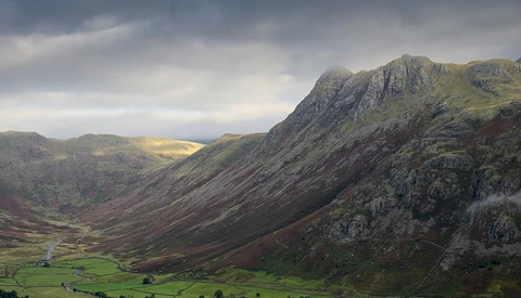 Dramatic mountain valley with steep rocky slopes, green fields below, and a small stream running through the landscape.