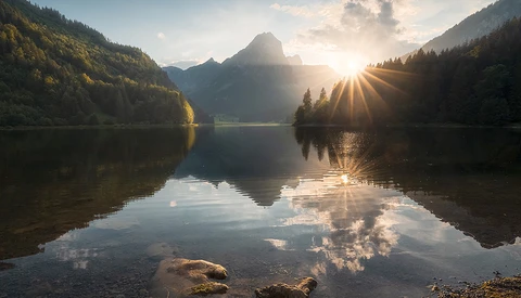 Mountain lake at sunrise with forested slopes and perfect mirror reflection in still water.