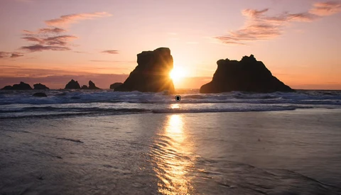 Sun setting between two large sea stacks silhouetted against golden sky and ocean.