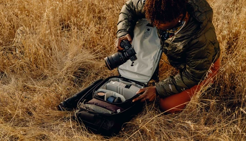 Photographer crouching in dry grass, adjusting a camera backpack with a large telephoto lens visible.