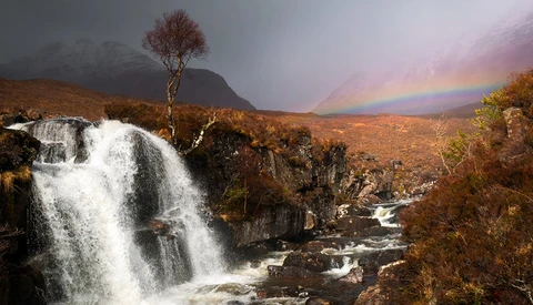 Waterfall cascading over rocky terrain with rainbow arcing across stormy sky and mountains.