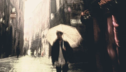 Figure holding white umbrella walking alone down a narrow, wet urban street.
