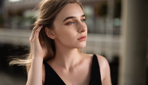 Woman in black tank top looking away, hand near ear, soft natural lighting from the side.