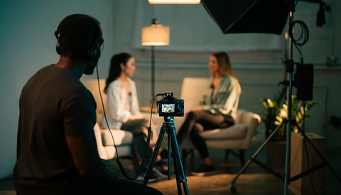 Camera on tripod in foreground during a studio interview setup with two people seated in background.