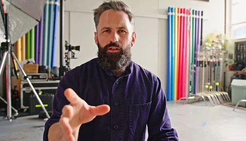 Bearded man in dark work shirt gesturing while speaking in a studio with colorful background rolls and lighting equipment.