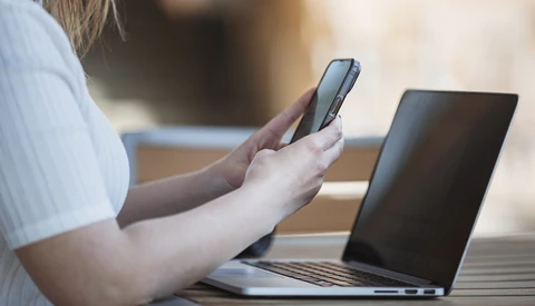 Person holding a smartphone while working at a laptop outdoors in natural daylight.