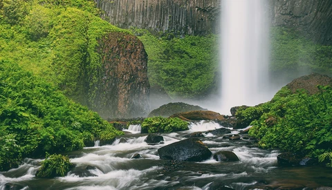 Waterfall cascading into a rocky stream surrounded by moss-covered boulders and lush green forest.