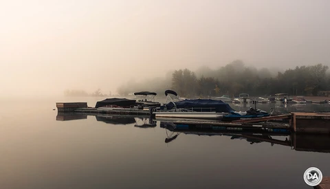 Moored boats reflected in still water at dawn with forested shoreline shrouded in fog.