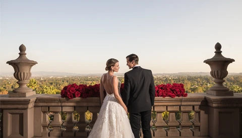 Bride and groom viewing expansive landscape from stone balustrade with ornamental urns and red floral arrangements.