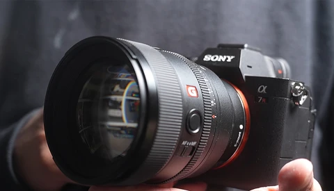 Sony mirrorless camera with telephoto lens mounted, held in hands against blurred gray background.