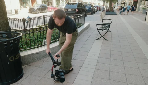 Photographer bending down to adjust a camera mounted on a skateboard on an urban sidewalk.