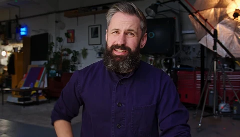 Bearded man in dark purple work shirt smiling in a workshop studio setting.