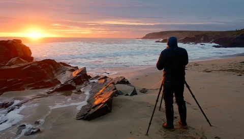 Photographer on a beach with tripod watching golden sunrise over ocean with rocky outcrops.