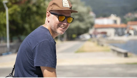 Man wearing a brown cap and yellow-tinted sunglasses smiles at the camera outdoors.