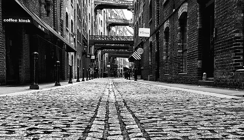 Cobblestone alley lined with brick buildings and overhead walkways, converging toward distant street.
