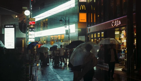 Rainy urban street at night with pedestrians holding umbrellas beneath illuminated storefronts and signs.