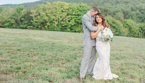 Bride and groom embracing in an open field with forested hills in the background.