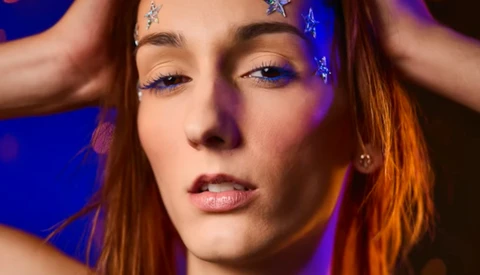 Woman with reddish-brown hair and blue eyeshadow against purple-blue lighting.