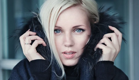 Portrait of a woman with platinum blonde hair and dark eye makeup holding black feathered props near her face.