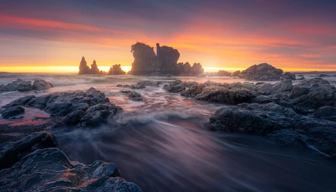 Dramatic coastal landscape with rocky outcrops and sea stacks silhouetted against a golden sunset.