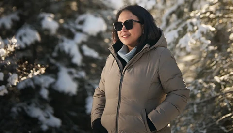 Woman in beige puffer jacket and sunglasses standing in snowy forest with evergreen trees.
