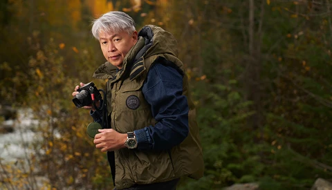 Photographer with gray hair holding a camera in an autumn forest setting.