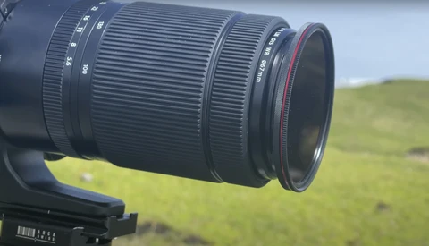 Telephoto zoom lens mounted on camera body with grassy hillside in soft focus background.