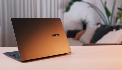 ASUS laptop with copper-colored lid angled on desk, blurred background with potted plant and desk accessories.