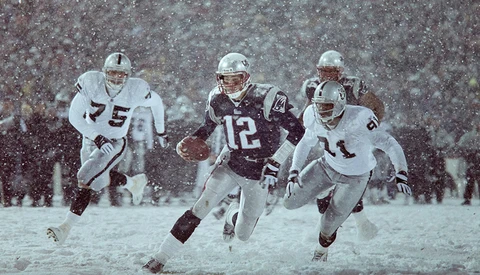 Four football players in a heavy snowstorm, two wearing white uniforms and two in dark uniforms with numbers visible.
