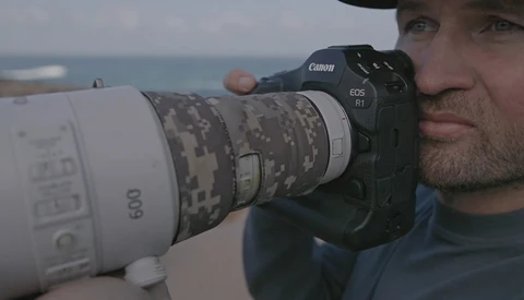 Photographer holding Canon EOS R1 with white telephoto lens featuring digital camouflage pattern.