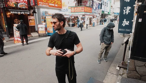 Man in black shirt and sunglasses holding a camera on a busy Japanese street lined with shops.
