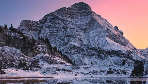 Snow-covered mountain peak reflected in a partially frozen lake at sunset, with evergreen trees along the shoreline.