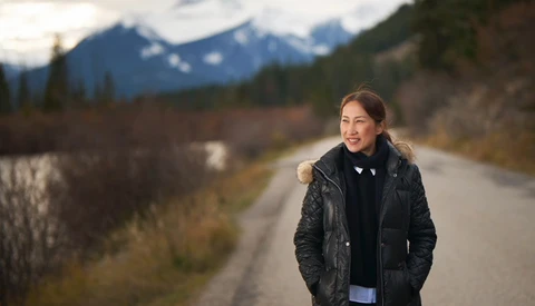 Woman in black quilted jacket smiling at camera on rural road with snow-capped mountains and river in background.
