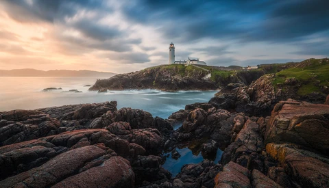 Lighthouse on rocky headland with turbulent ocean and dramatic cloudy sky at dusk.