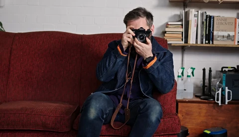 Photographer holding vintage camera to face while sitting on red couch in home studio.