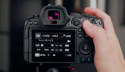 Photographer's hand holding a DSLR camera displaying exposure settings on the rear LCD screen.