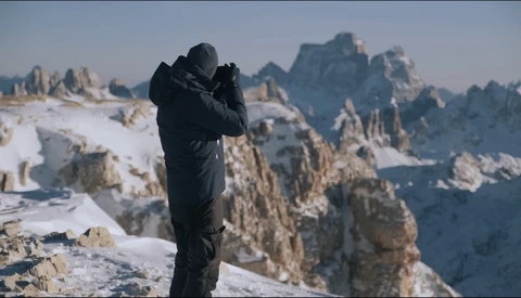 Photographer in black jacket holding camera to face on snowy mountain peak with dramatic rocky peaks in background.