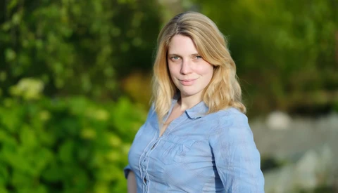 Woman with blonde shoulder-length hair wearing a light blue button-up shirt, photographed outdoors with soft natural lighting.