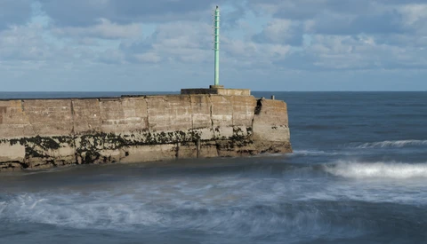Weathered stone pier or breakwater extending into rough ocean waves under partly cloudy sky.
