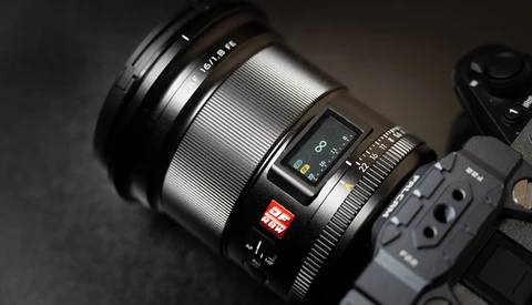 Close-up of a mirrorless camera lens with manual focus ring and autofocus controls mounted on a camera body against a dark surface.
