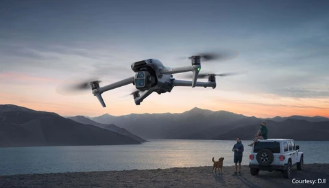 Drone hovering above a beach at sunset with people and a vehicle below.