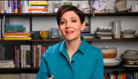 Woman in turquoise shirt speaking in front of shelving with books and kitchenware.