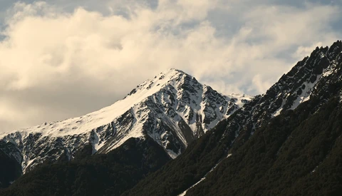 Snow-capped mountain peak with forested slopes under partly cloudy sky.
