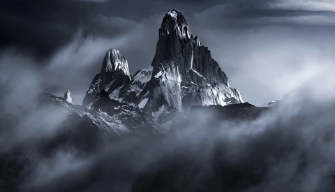 Snow-covered jagged mountain peaks rising through thick clouds under a dramatic gray sky.