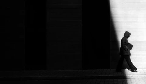 Silhouetted figure in dark clothing walking across a stark white floor against a black wall.